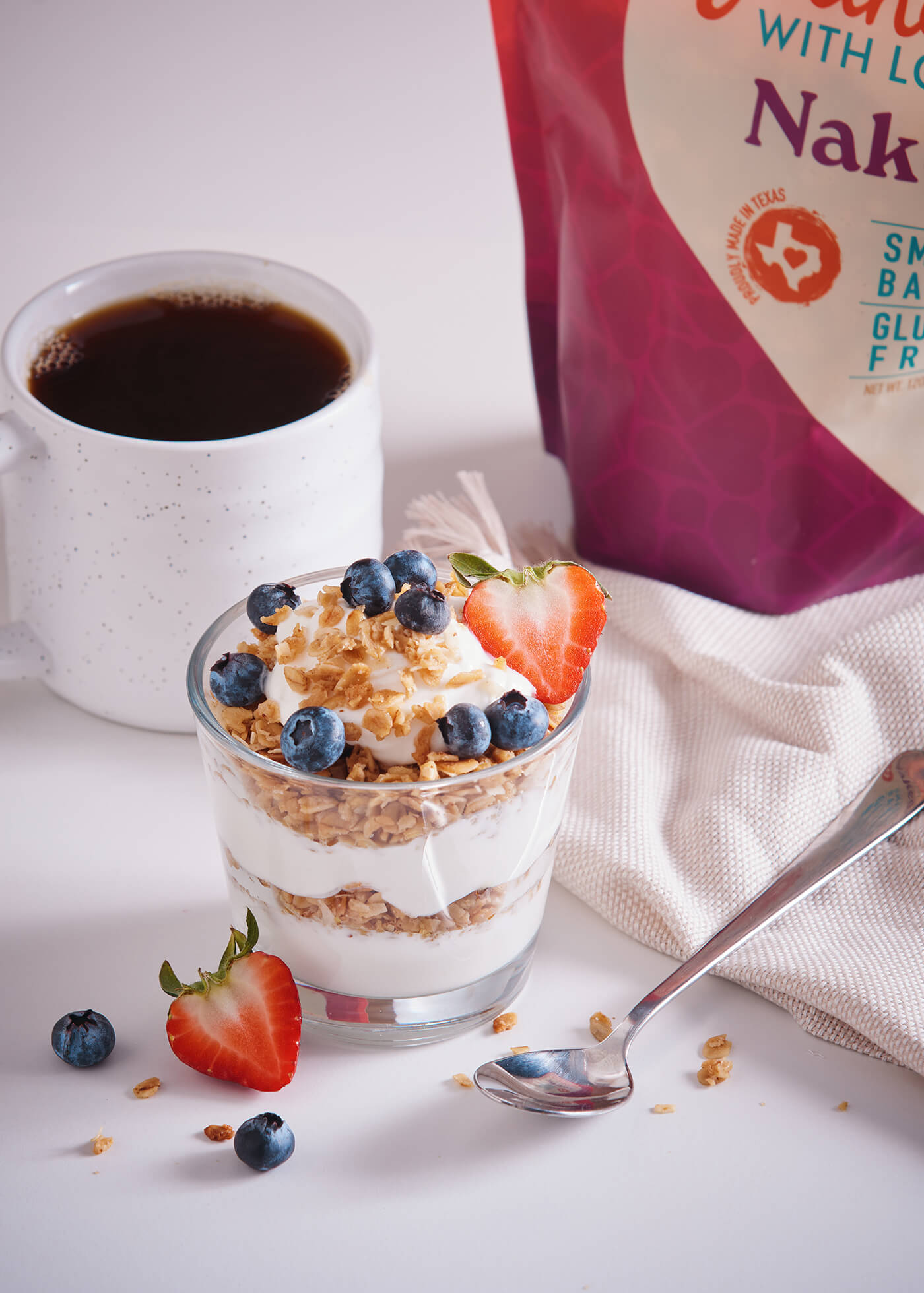 Glass dessert with layers of cream, fruit, and granola next to a cup of coffee and a bag of cereal.